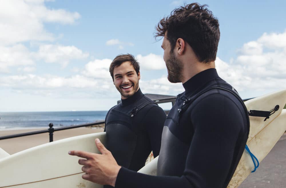 Gabriel Vigil and James walk towards the beach with surfboards on a sunny day.