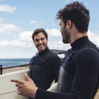 Gabriel Vigil and James walk towards the beach with surfboards on a sunny day.