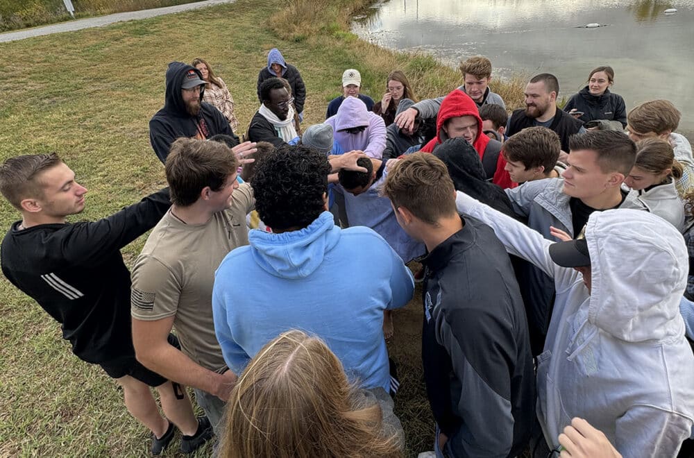 College students in a circle praying over a friend by a pond under an overcast sky.