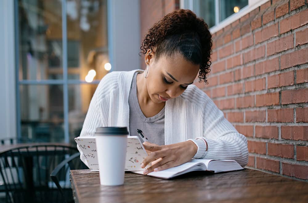 A mixed race women reads a large Bible / textbook while seated at table outdoors at a coffee shop.
