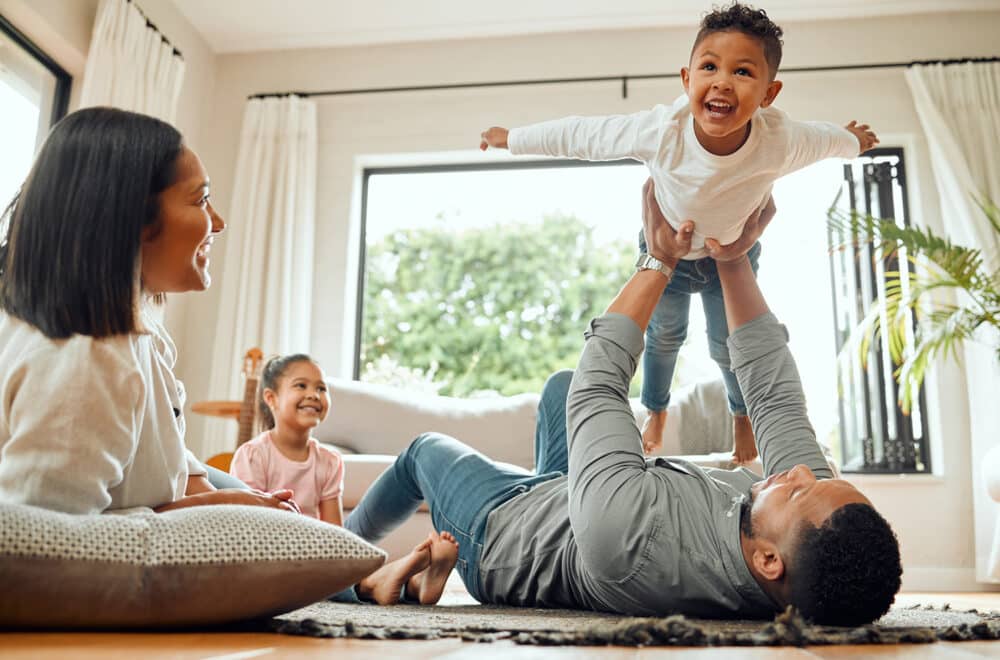 A family lays on the ground together while the father lifts up his son towards the ceiling.