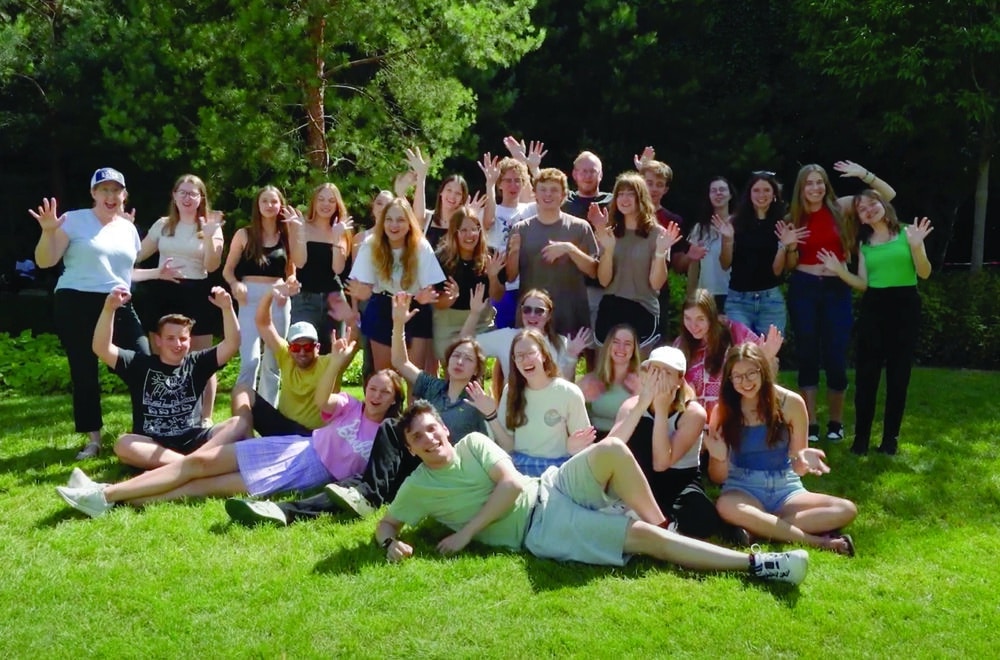 A large group of young adults poses together on a sunny lawn, smiling and waving enthusiastically at the camera.