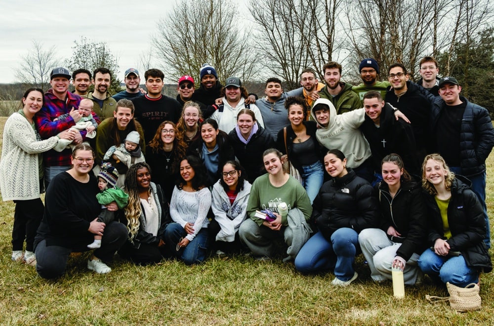 A large group of young adults and a few small children pose together outdoors on a cloudy day, smiling warmly at the camera. They are bundled up in winter and fall clothing, standing on a grassy field with bare trees in the background.