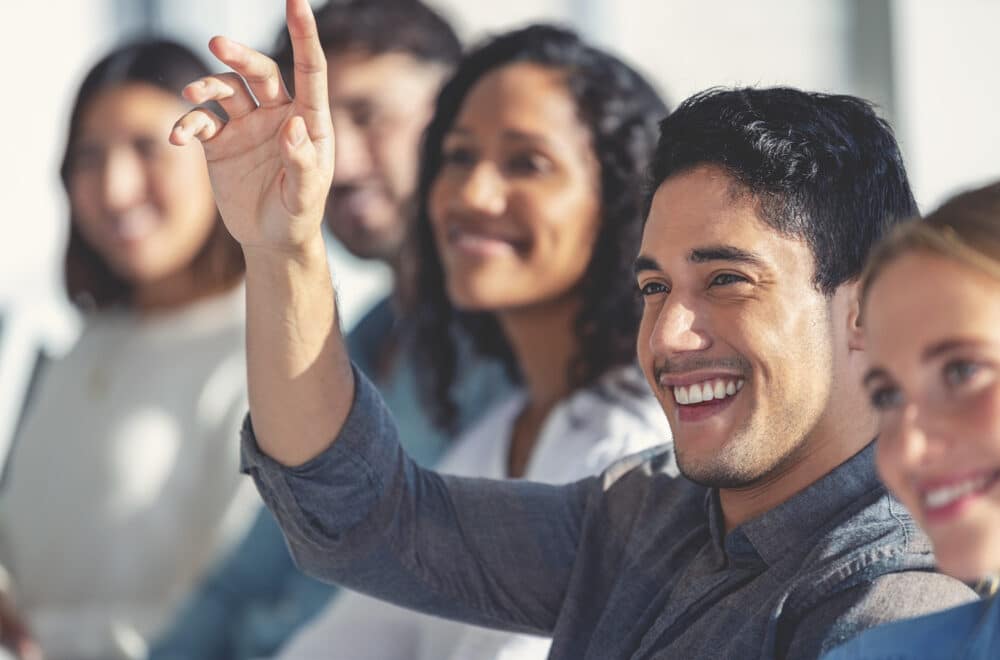 A close up image of a smiling man raising his hand to ask a question