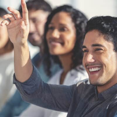 A close up image of a smiling man raising his hand to ask a question