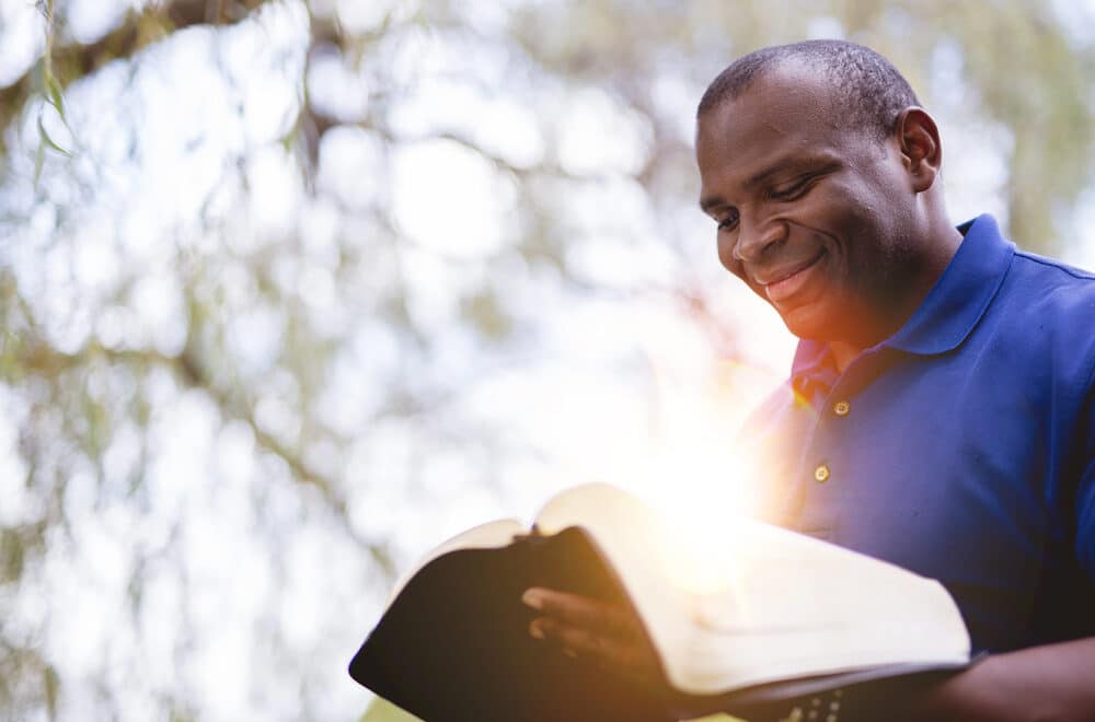 A man wearing a blue polo shirt smiles as he reads an open Bible outdoors. Sunlight shines brightly from the pages, and a soft-focus background of trees creates a peaceful atmosphere.
