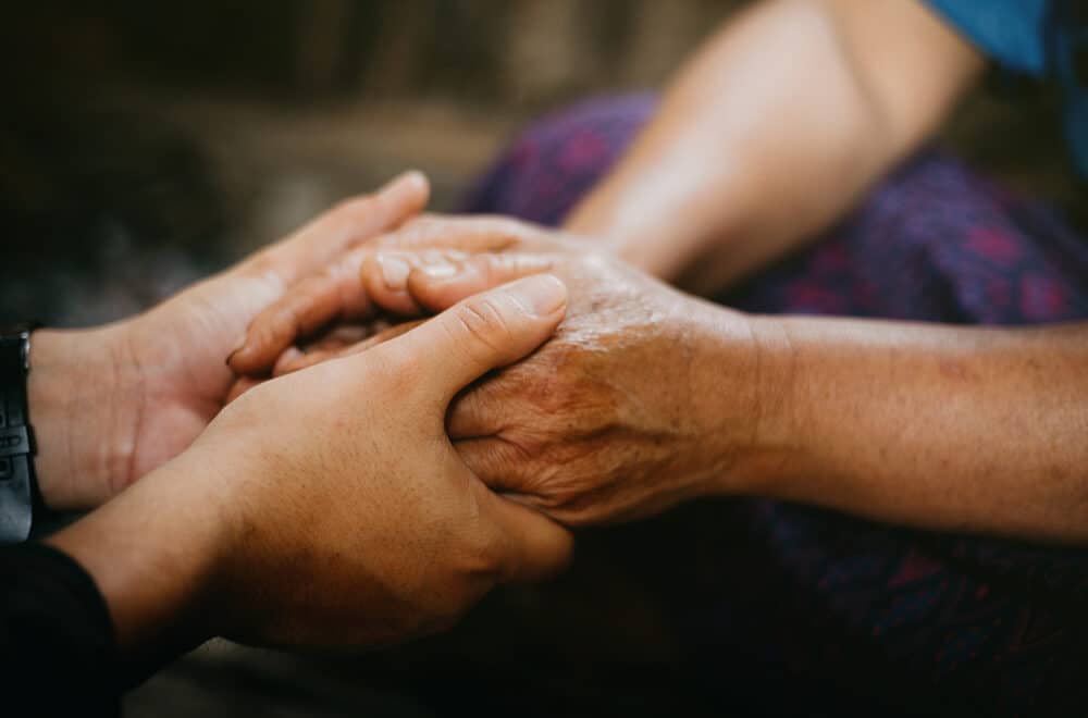 Two people gently hold hands in a comforting gesture. One pair of hands appears older and weathered, while the other pair is younger.