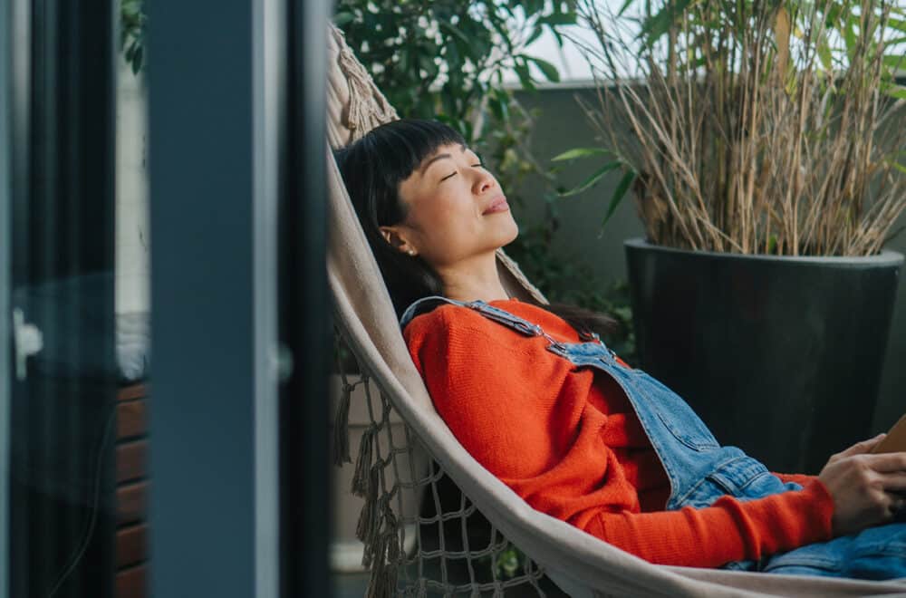 A woman relaxes in a hammock on a balcony surrounded by potted plants. She is wearing a red sweater and denim overalls, with her eyes closed and a peaceful expression. She holds a closed book in one hand, and soft natural light fills the scene.