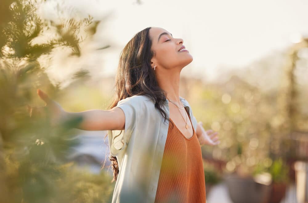 Young latin woman with arms outstretched breathing in fresh air during sunrise at the balcony. Healthy girl enjoying nature while meditating during morning with open arms and closed eyes. Mindful woman enjoying morning ritual while relaxing in outdoor park.