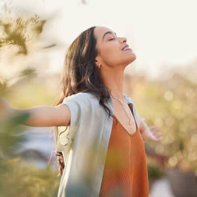 Young latin woman with arms outstretched breathing in fresh air during sunrise at the balcony. Healthy girl enjoying nature while meditating during morning with open arms and closed eyes. Mindful woman enjoying morning ritual while relaxing in outdoor park.