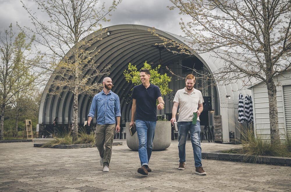 Three men walk outdoors in front of a modern, arched metal building, each holding a Bible and smiling in conversation, surrounded by leafless trees and cloudy skies.