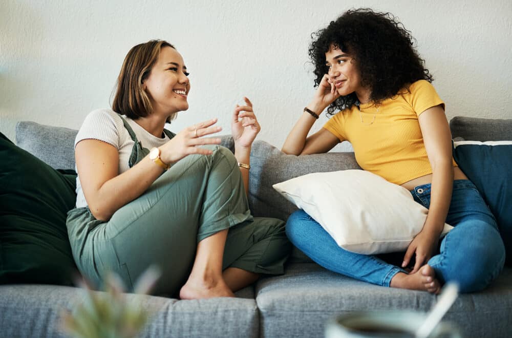 Two women talking in a home on a couch, smiling while in conversation.