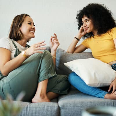 Two women talking in a home on a couch, smiling while in conversation.