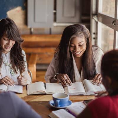 Four women sitting at a wooden table in a café, smiling and reading open Bibles together during a small group study.