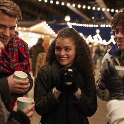 A group of friends smiles and talks while holding hot drinks at an outdoor market lit by string lights at night.