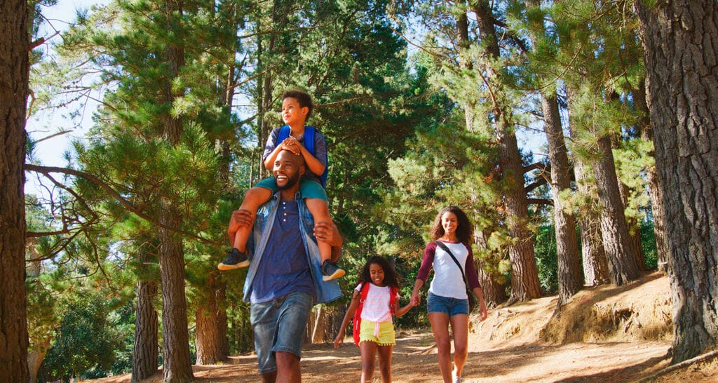 A family walks through a forest trail together, with the father carrying a child on his shoulders and the mother and daughter walking beside them.