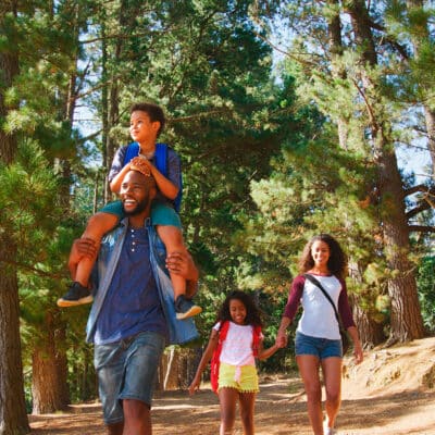 A family walks through a forest trail together, with the father carrying a child on his shoulders and the mother and daughter walking beside them.