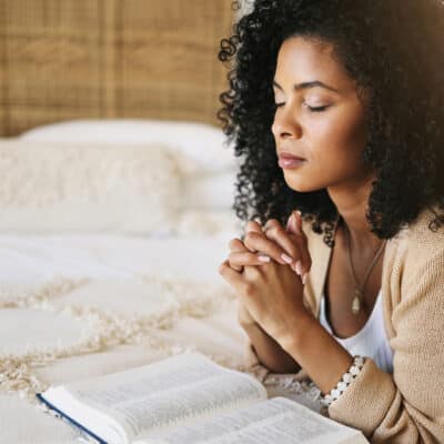 A woman sits on a bed with her hands clasped in prayer, eyes closed, beside an open Bible resting on the blanket.