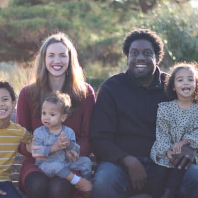 A family of five sits together on an outdoor bench, smiling and laughing in the sunlight.