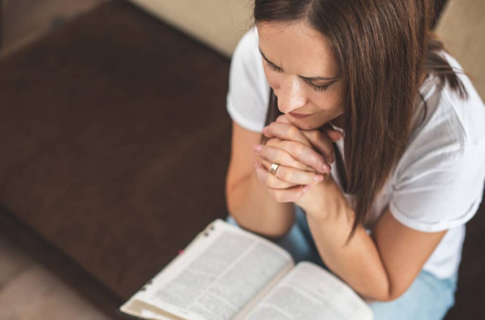A woman sits with her hands clasped in prayer, leaning over an open Bible in her lap.