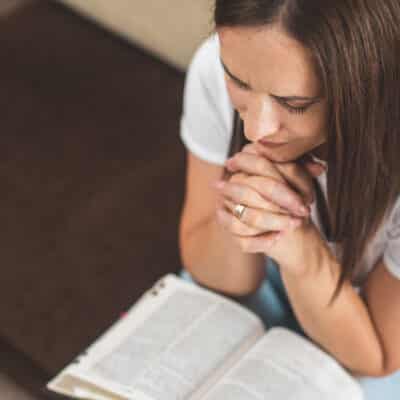 A woman sits with her hands clasped in prayer, leaning over an open Bible in her lap.