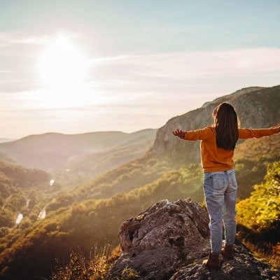 Person standing on a rocky overlook with arms outstretched, facing a sunlit mountain landscape with autumn-colored trees.