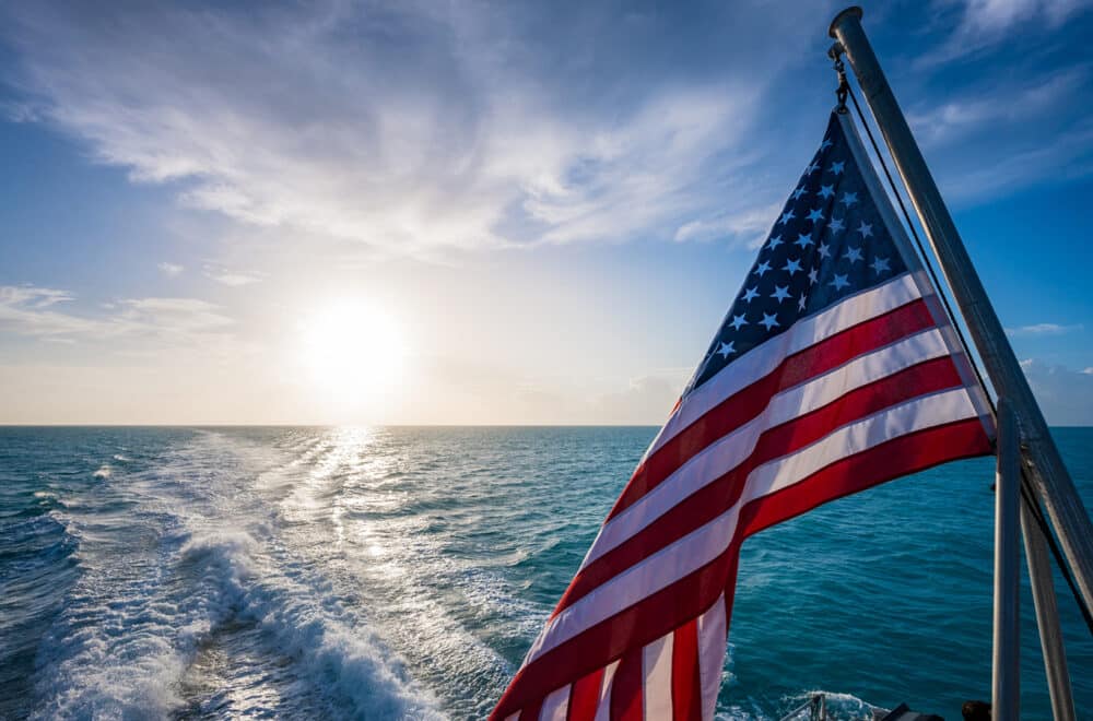 An American flag waves off the back of a boat with the wake from the boat in the ocean behind it.