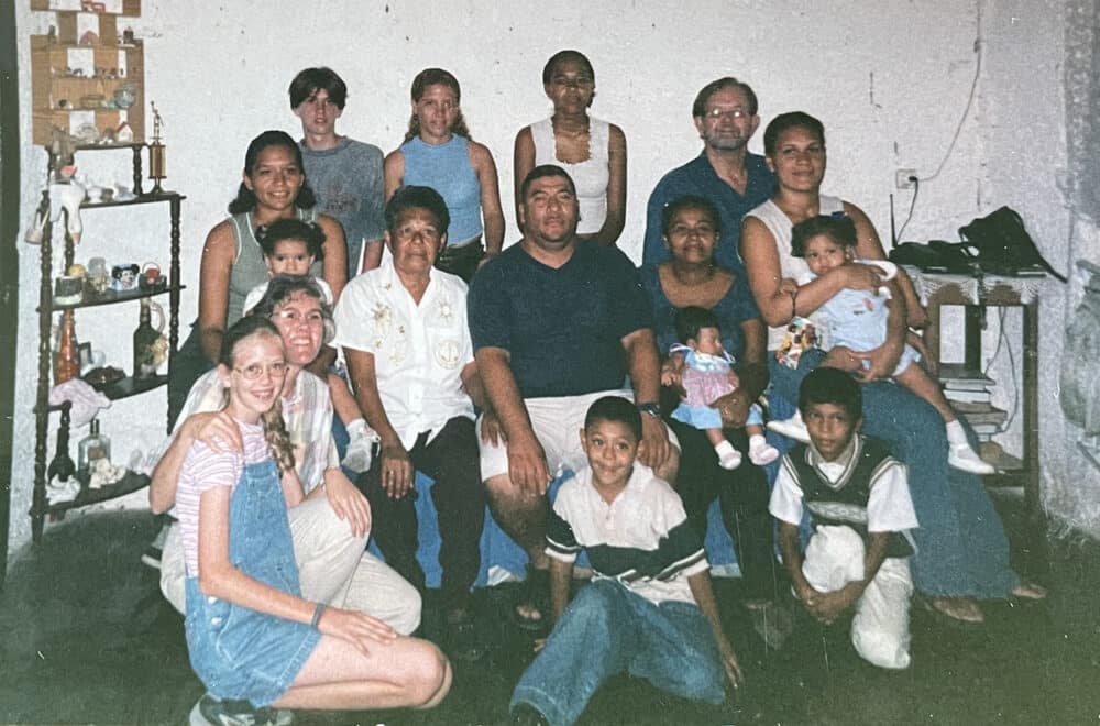 Multi-generational family group gathered indoors, posing together in a living room with adults, children, and babies.