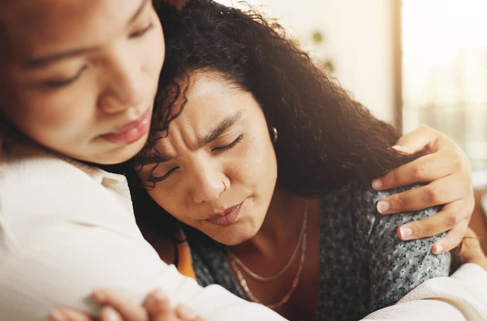 Two women sitting close together, one person gently embracing the other in a moment of comfort and support.