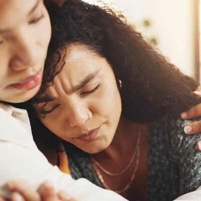 Two women sitting close together, one person gently embracing the other in a moment of comfort and support.