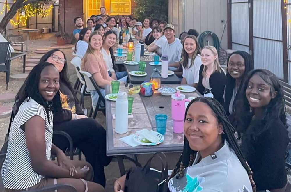 Large group of adults seated at a long outdoor table, smiling and sharing a meal together in a backyard setting with string lights.