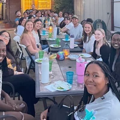 Large group of adults seated at a long outdoor table, smiling and sharing a meal together in a backyard setting with string lights.