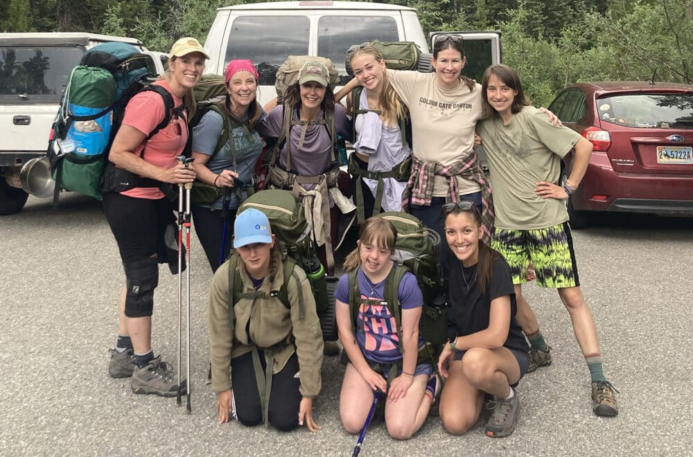 A group of nine hikers pose together in a forest parking area, smiling in front of a white van. They are wearing backpacks and outdoor gear, with some holding trekking poles.