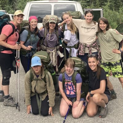 A group of nine hikers pose together in a forest parking area, smiling in front of a white van. They are wearing backpacks and outdoor gear, with some holding trekking poles.