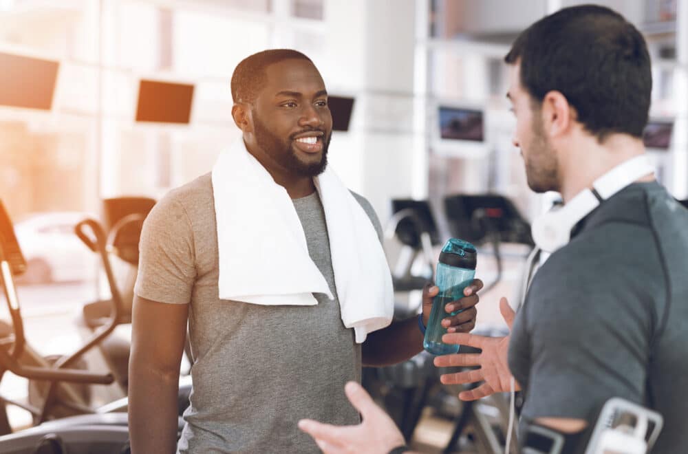 Two men talking in a gym, one holding a water bottle and wearing a towel around his neck after a workout.