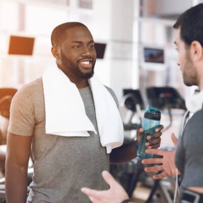 Two men talking in a gym, one holding a water bottle and wearing a towel around his neck after a workout.