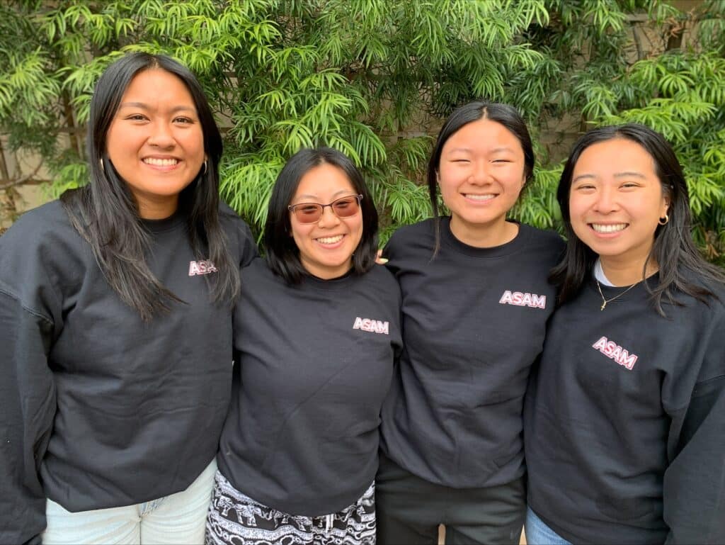 Four smiling women wearing matching black “ASAM” sweatshirts stand close together with their arms around each other in front of lush green foliage.