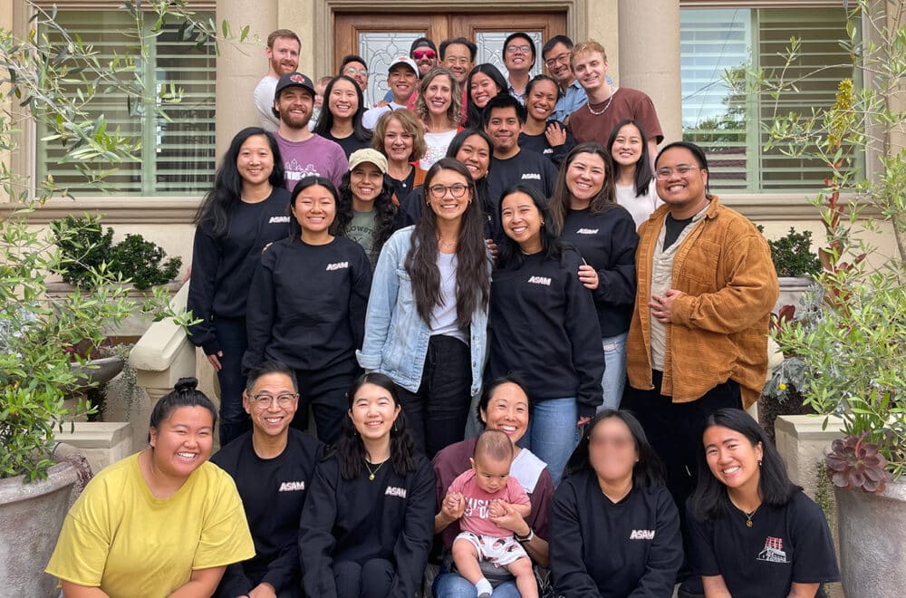 A large, diverse group of adults and one baby smiling and posing together on the front steps of a house.