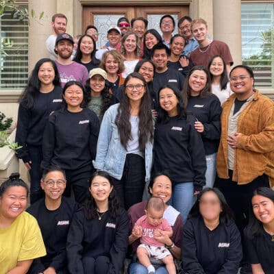 A large, diverse group of adults and one baby smiling and posing together on the front steps of a house.