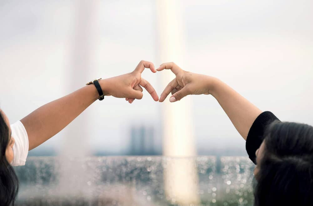 Two hands forming a heart shape together against a blurred outdoor background, symbolizing love and connection.