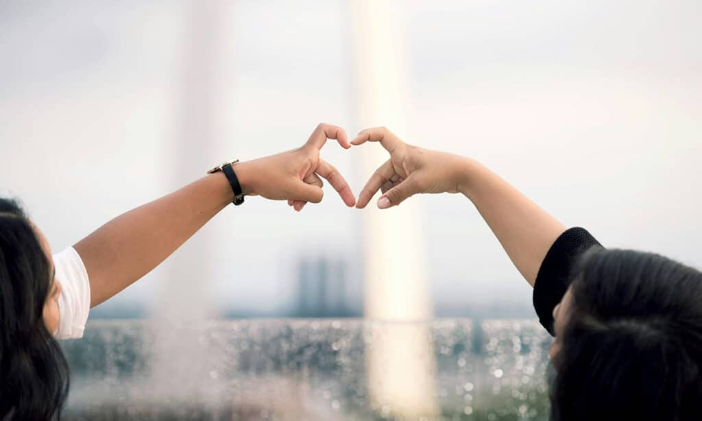Two hands forming a heart shape together against a blurred outdoor background, symbolizing love and connection.