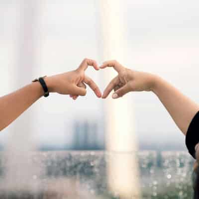 Two hands forming a heart shape together against a blurred outdoor background, symbolizing love and connection.