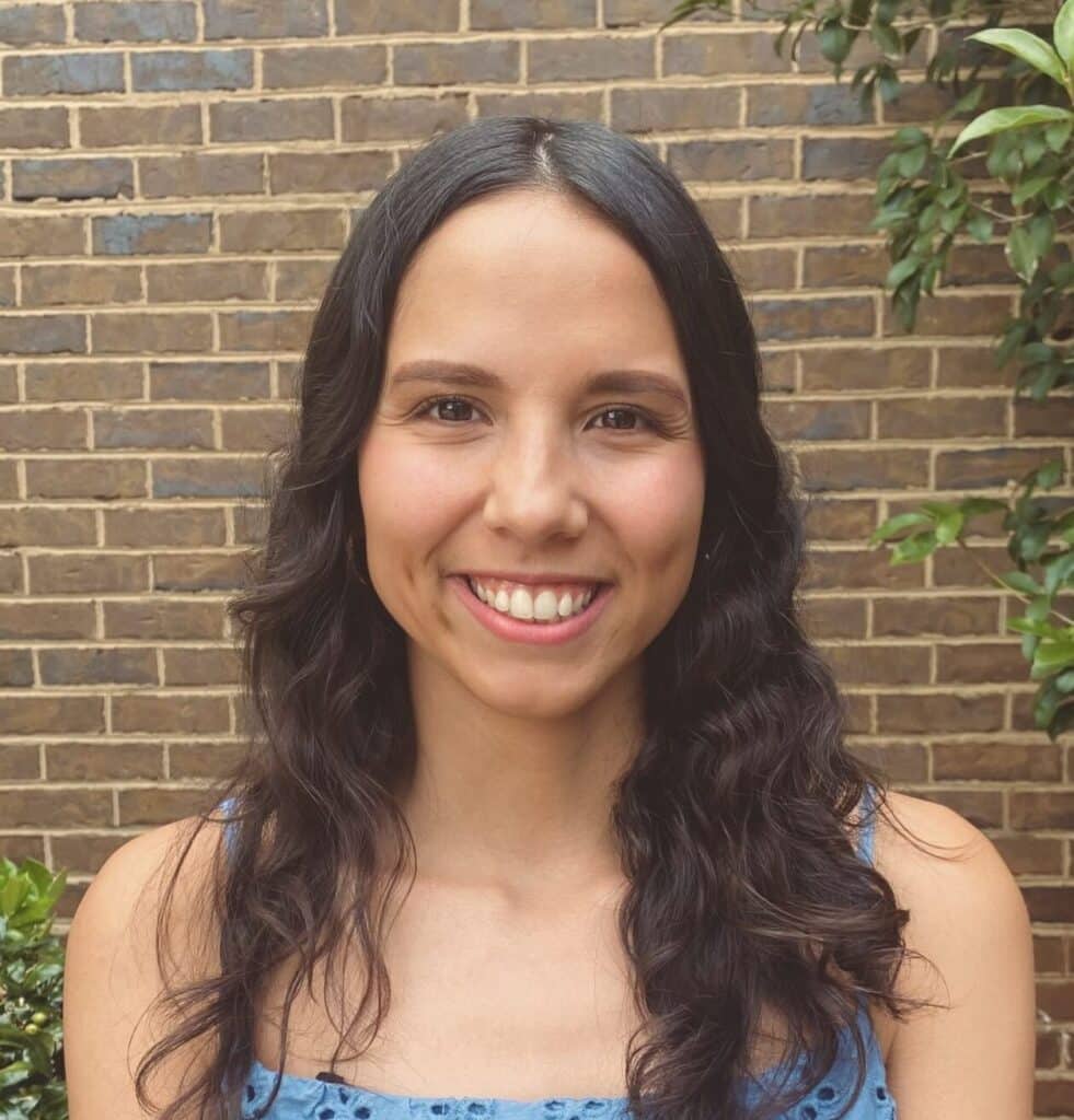 Smiling woman with long wavy dark hair stands in front of a brick wall outdoors.