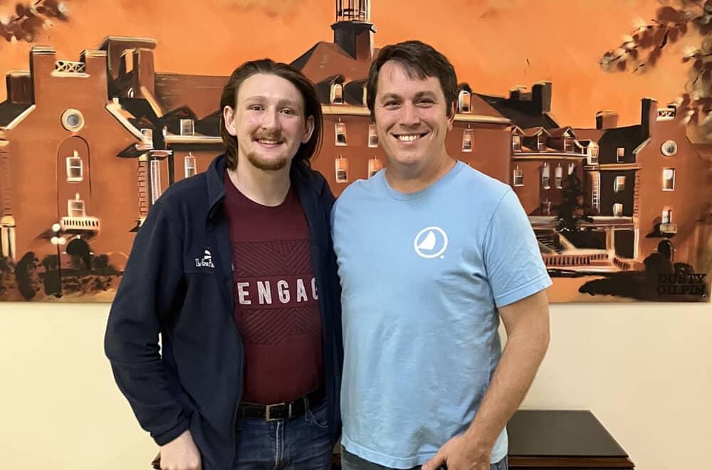 Two men posing together indoors, arms around each other, smiling in front of an orange mural depicting buildings and a tower.