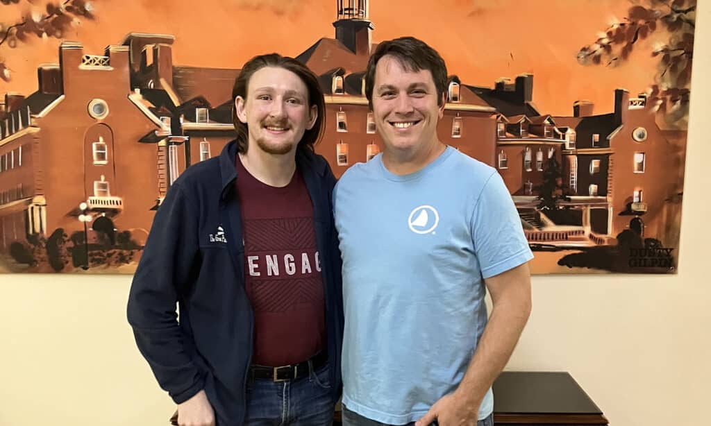 Two men posing together indoors, arms around each other, smiling in front of an orange mural depicting buildings and a tower.