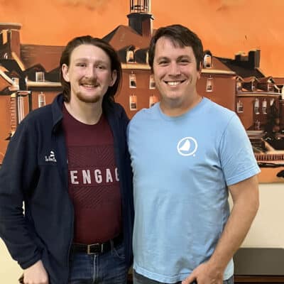 Two men posing together indoors, arms around each other, smiling in front of an orange mural depicting buildings and a tower.