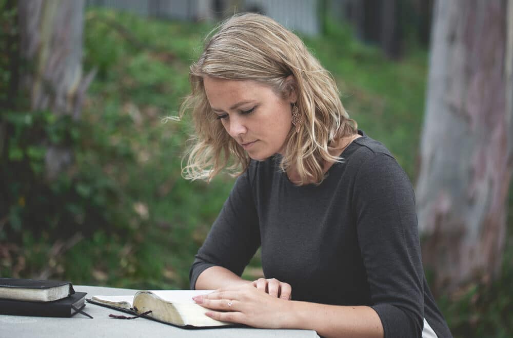 A person sitting at a round concrete table outdoors, reading an open book with thin pages, likely a Bible, while another closed book rests nearby.