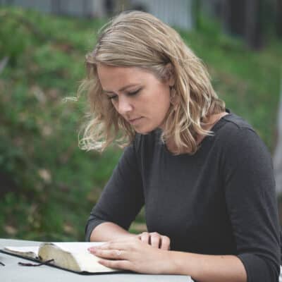 A person sitting at a round concrete table outdoors, reading an open book with thin pages, likely a Bible, while another closed book rests nearby.