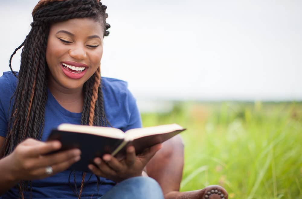 Smiling woman with braided hair reading an open book outdoors, with a blurred green background and decorative circular pattern overlay on the right side of the image.