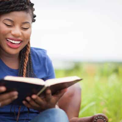Smiling woman with braided hair reading an open book outdoors, with a blurred green background and decorative circular pattern overlay on the right side of the image.